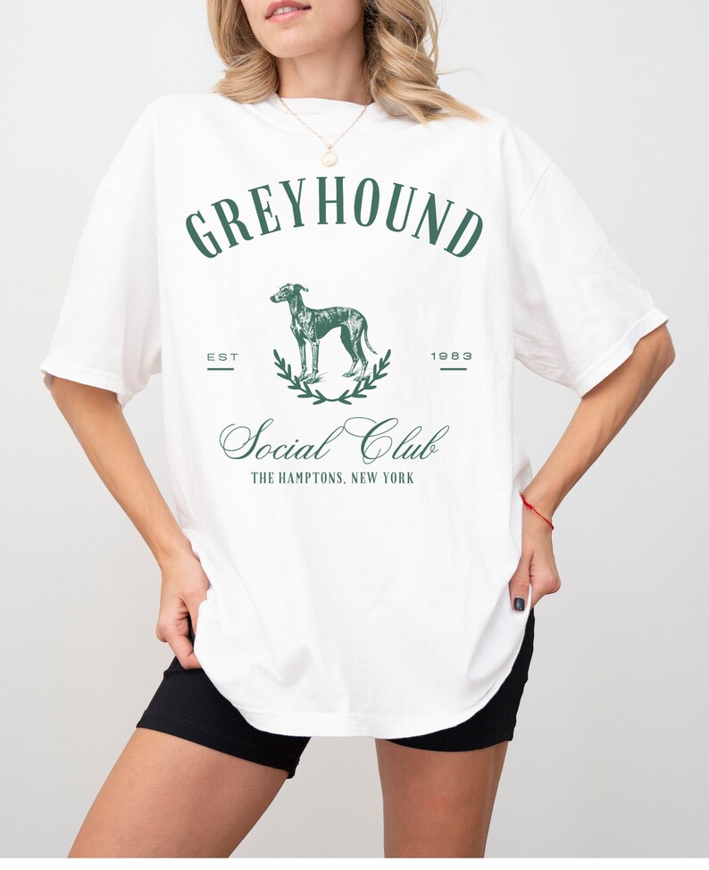 以下が含まれることがあります： グレーのグレイハウンド犬、月桂樹の輪、そして「Greyhound Social Club The Hamptons, New York」というテキストと「Est 1983」というテキストが書かれたグリーンのグラフィックプリントが施された、白のオーバーサイズTシャツです。
