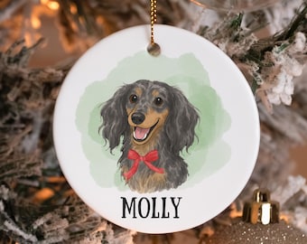 May include: A white ceramic Christmas ornament featuring a long-haired dachshund illustration with a red bow tie. The dog has black and brown fur and a happy expression. The name "MOLLY" is printed in black below the dog.