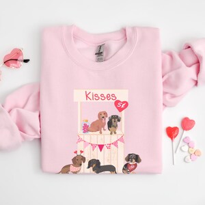 Puede incluir: Sudadera rosa con un gr&aacute;fico de un puesto de besos de dachshund con el texto "Kisses 5&cent;".