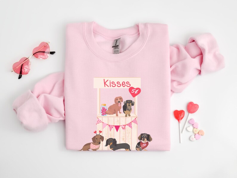 Puede incluir: Sudadera rosa con un gr&aacute;fico de un puesto de besos de dachshund con el texto "Kisses 5&cent;".