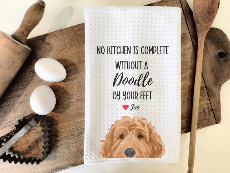 a kitchen towel with a dog illustration and a humorous message about cooking, placed on a wooden cutting board alongside kitchen utensils.