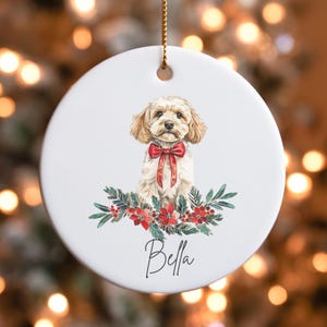 May include: A white ceramic Christmas ornament featuring a watercolor illustration of a small, light brown dog wearing a red bow. The dog is surrounded by green foliage and red berries. The ornament is personalized with the name "Bella".