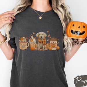 May include: A dark gray t-shirt with a graphic of a golden retriever puppy wearing a scarf, surrounded by fall-themed imagery, including coffee cups, pumpkins, and leaves. The text "Pumpkin Spice" is on one of the coffee cups.