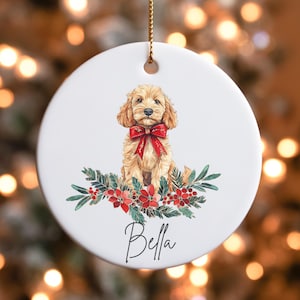 May include: A round, white ceramic Christmas ornament featuring a golden-colored dog with a red bow, sitting above a floral arrangement of red flowers and green leaves. The name "Bella" is written in cursive below the dog.