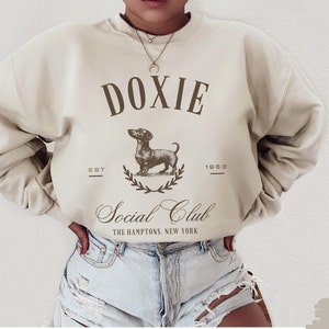 May include: A beige sweatshirt with a brown graphic of a dachshund and the text "DOXIE Social Club THE HAMPTONS, NEW YORK EST 1983"