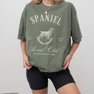 May include: A moss green t-shirt with a white graphic of a spaniel dog and the text "Spaniel Social Club The Hamptons, New York" with the text "Est 1850" above the dog.