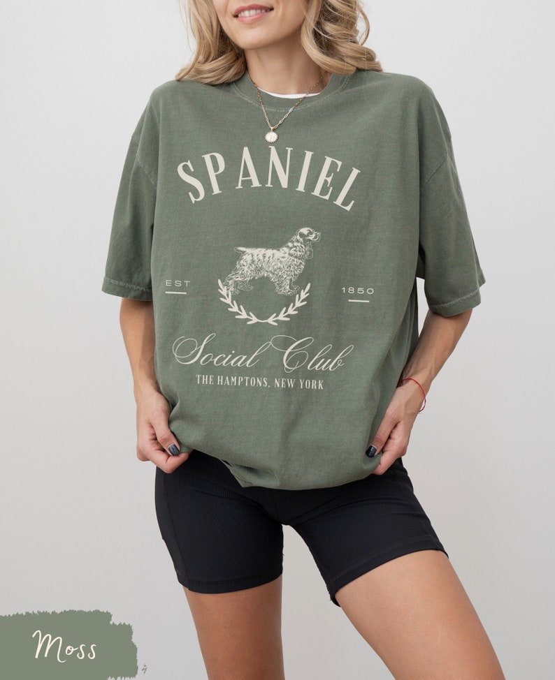 May include: A moss green t-shirt with a white graphic of a spaniel dog and the text "Spaniel Social Club The Hamptons, New York" with the text "Est 1850" above the dog.