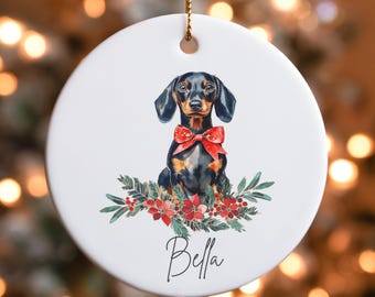 May include: A white ceramic Christmas ornament featuring a black and tan dachshund wearing a red bow. The dog is surrounded by green holly and red berries. The ornament is personalized with the name "Bella".