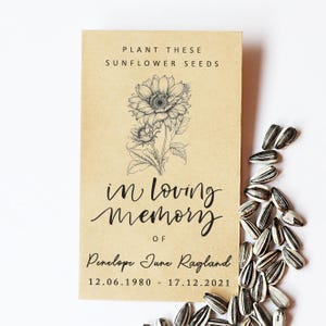 Op de afbeelding: Een bruin zaadzakje met de tekst "Plant deze zonnebloempitten" en een gedetailleerde illustratie van een zonnebloem. De tekst "In Loving Memory Of Penelope June Ragland 12.06.1980 - 17.12.2021" is ook aanwezig. Zonnebloempitten zijn verspreid.