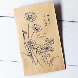 Könnte beinhalten: Ein beigefarbenes Notizbuch mit einer schwarzen Blumenillustration und dem Text "24 08 23" und "MITCH CASEY". Das Design des Notizbuchs ist schlicht und elegant, mit Fokus auf die botanische Illustration.