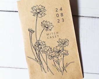 May include: A tan-colored notebook featuring a black floral illustration and the text "24 08 23" and "MITCH CASEY". The notebook has a simple, elegant design with a focus on the botanical artwork.