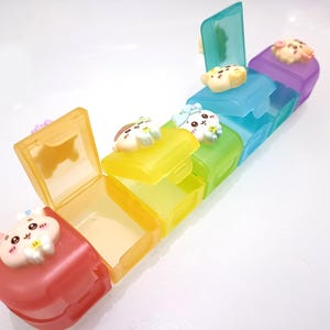 May include: A colorful, multi-compartment pill organizer with a hinged lid on each section. The sections are red, yellow, green, blue, and purple. Each lid is decorated with a cute cartoon animal.