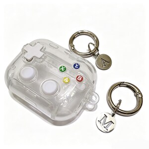May include: A clear protective case for wireless earbuds designed to resemble a video game controller, featuring a white directional pad, two white joysticks, and four colored buttons labeled X, Y, A, and B. Two silver keychains with letter charms A and M.