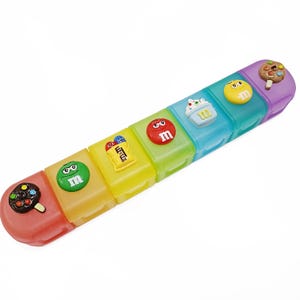 May include: A colorful, segmented pill organizer with seven compartments. Each compartment is a different color and has a candy-themed design on top, including ice cream, cupcakes, and M&M's candies. The organizer is designed to hold pills or small items.