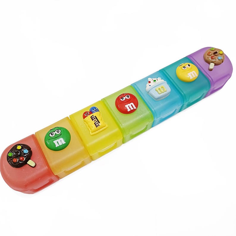 May include: A colorful, segmented pill organizer with seven compartments. Each compartment is a different color and has a candy-themed design on top, including ice cream, cupcakes, and M&M's candies. The organizer is designed to hold pills or small items.