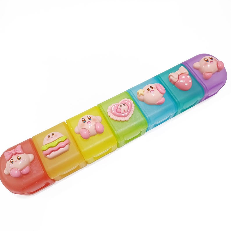 May include: A colorful pill organizer featuring six compartments in red, yellow, green, blue, and purple. Each compartment has a different pink Kirby character design on top. The organizer is designed to hold pills or small items.