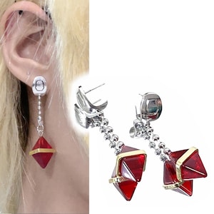 May include: Dangle earrings in silver, featuring a geometric design. Each earring has a silver top, a chain of small silver beads, and a red, diamond-shaped pendant with gold-coloured trim. The earrings are shown on a person's ear.