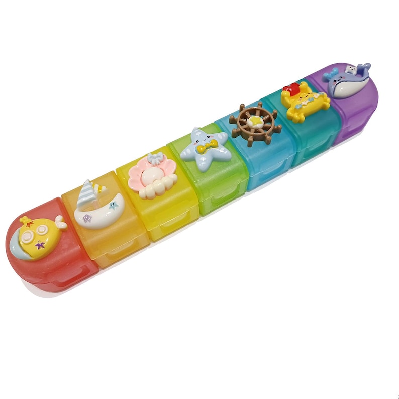 May include: A colorful, rectangular pill organizer with seven compartments, each a different color of the rainbow. Each compartment is decorated with a different nautical-themed charm, including a submarine, sailboat, starfish, and whale.