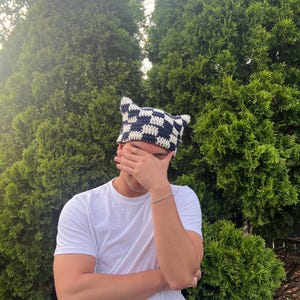 Puede incluir: Un sombrero de ganchillo hecho a mano con orejas de gato. El sombrero presenta un patrón de cuadros en azul marino y blanco. El sombrero lo lleva una persona que lleva una camiseta blanca.