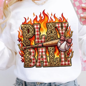 May include: White sweatshirt with a baseball-themed graphic. The design features the words "PLAY BALL" in leopard print and red plaid, with a baseball bat, ball, and flames. Includes horseshoes and stars.