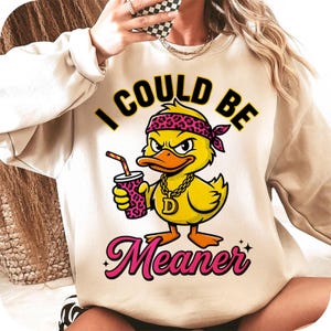 Könnte beinhalten: Beiges Sweatshirt mit einer Grafik einer gelben Ente mit rosa Bandana und goldener Kette, die ein Getränk hält. Der Text lautet "I COULD BE Meaner" in Schwarz und Pink.
