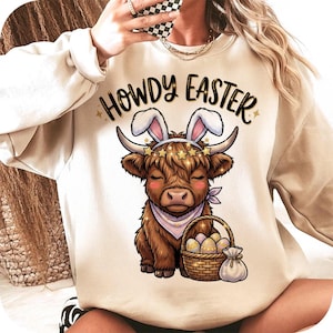 Puede incluir: Sudadera beige con el texto "HOWDY EASTER" sobre una vaca de las Tierras Altas de dibujos animados con orejas de conejo y un pañuelo. La vaca sostiene una cesta de huevos de Pascua. El diseño utiliza tonos marrones, rosas y dorados.
