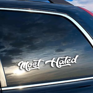 May include: White vinyl decal with the words "Most Hated" in a stylized script font. The decal is applied to the window of a dark-colored vehicle, with a cloudy sky visible through the glass.
