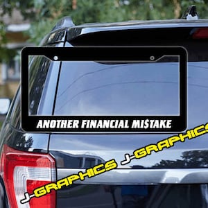 May include: Black license plate frame with the text "Another Financial Mistake" in white letters. The frame is attached to the back of a black SUV.