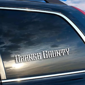 May include: White vinyl decal with the text "ORANGE COUNTY" in a decorative font, applied to a car window. The background shows a cloudy sky with a hint of sunlight.