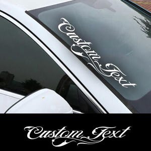 May include: White vinyl decal with the text "Custom Text" in a stylized font. The decal is applied to the windshield of a car.