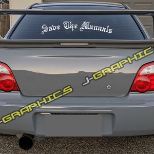 May include: A gray Subaru Impreza with a rear spoiler and a white vinyl decal that reads "Save The Manuals" on the rear window. The car has a black exhaust pipe and red taillights.