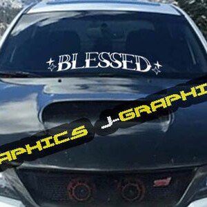 May include: A white car with a black hood and a windshield decal that says "BLESSED" with stars on either side.