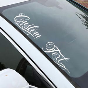 May include: White vinyl decal applied to a car windshield. The decal features the words "Custom Text" in an elegant, cursive font. The car is white, and the windshield reflects the sky and surrounding environment.