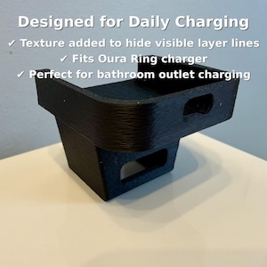 May include: A black 3D-printed charging stand for an Oura Ring. The stand is designed for daily charging and features a textured surface to hide layer lines. Text on the image reads "Designed for Daily Charging".