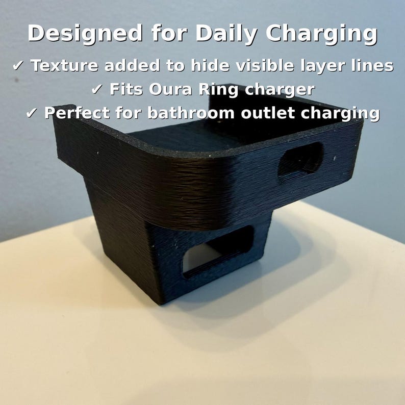 May include: A black 3D-printed charging stand for an Oura Ring. The stand is designed for daily charging and features a textured surface to hide layer lines. Text on the image reads "Designed for Daily Charging".