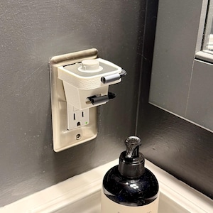May include: A white and gray electrical outlet cover with a built-in device, installed on a gray wall. The device has a small, rectangular shape with a circular top and two black handles. A black soap dispenser sits below.