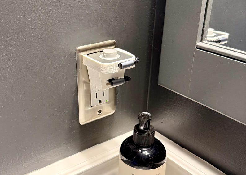 May include: A white and gray electrical outlet cover with a built-in device, installed on a gray wall. The device has a small, rectangular shape with a circular top and two black handles. A black soap dispenser sits below.