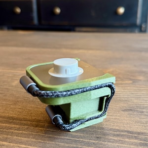 May include: A small, olive green electronic device with a white circular top and a black braided cord. The device has a rectangular shape with rounded edges and is resting on a wooden surface.
