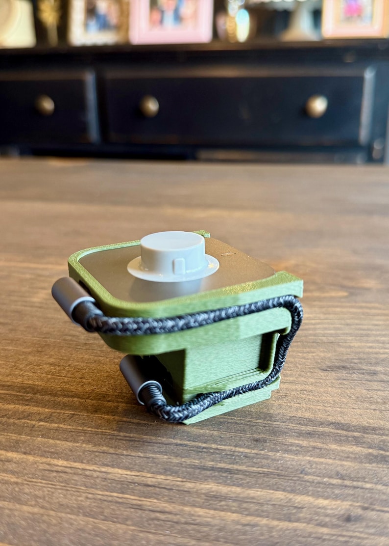 May include: A small, olive green electronic device with a white circular top and a black braided cord. The device has a rectangular shape with rounded edges and is resting on a wooden surface.