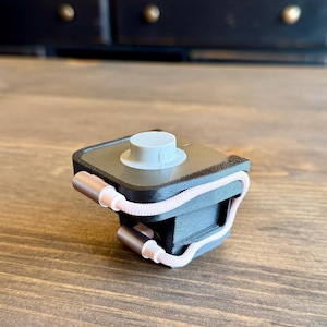 May include: A black and gray device with a white top and a pink and white cord. The device has two rose gold colored metal ends. The device is sitting on a wooden surface.