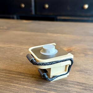 May include: A small, beige and gray device with a circular top and a black braided cord. The device has two gray cylindrical handles and sits on a wooden surface.