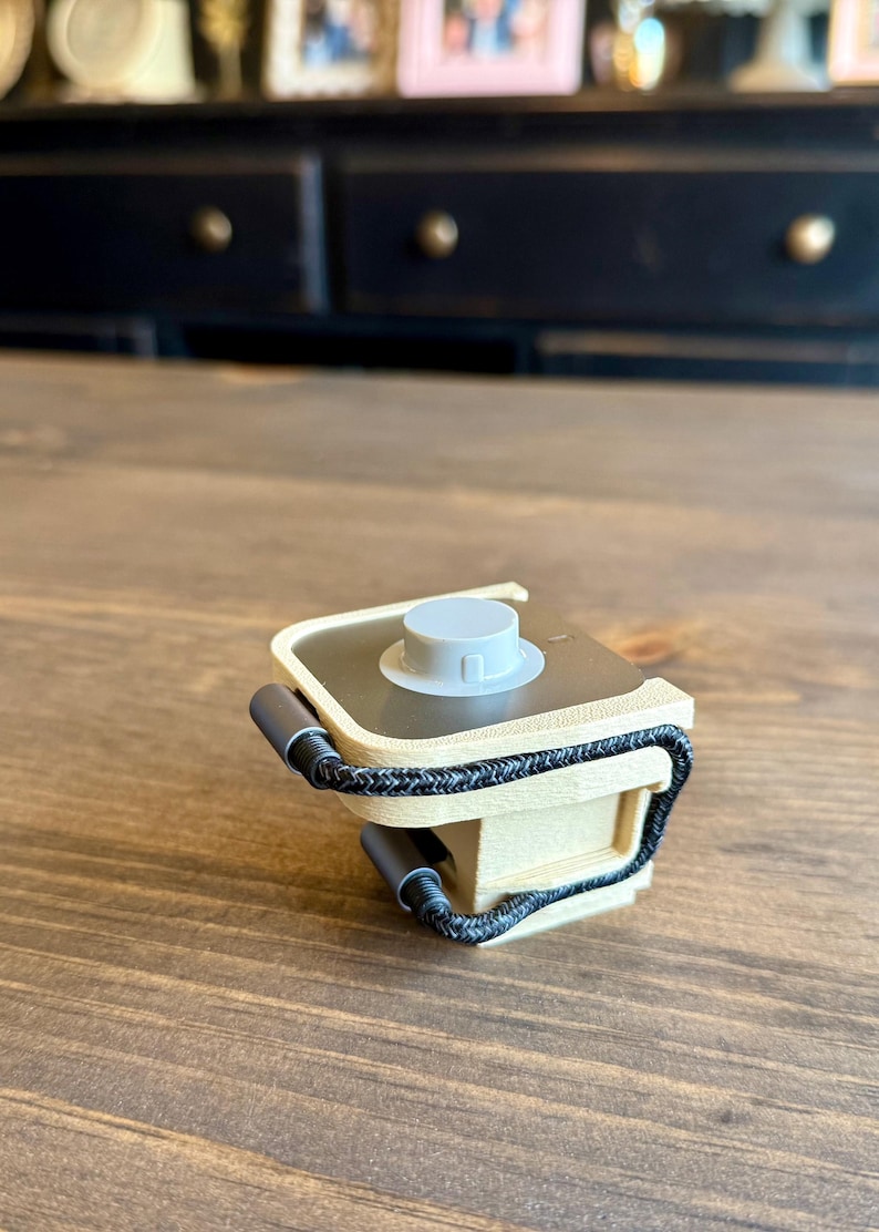 May include: A small, beige and gray device with a circular top and a black braided cord. The device has two gray cylindrical handles and sits on a wooden surface.