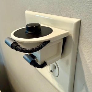 May include: A white and black charging station accessory, mounted on a wall outlet. The station features a black top, a white base, and two gray charging cables with a braided black cord. The outlet cover is white.