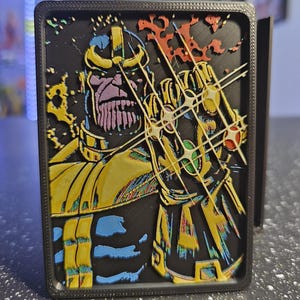 May include: A rectangular 3D-printed wall art piece featuring a layered relief of the character Thanos. The design displays Thanos in purple, yellow, and blue, wearing a gold gauntlet with colored stones, all set against a solid black background.
