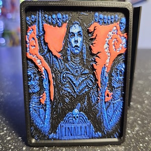 May include: A black rectangular relief sculpture featuring a central blue figure with outstretched arms, surrounded by red accents and skulls. The word "INALLA" is at the bottom. The artwork is framed in a black border.