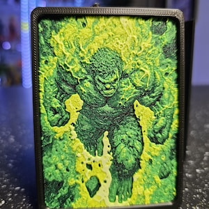 May include: A rectangular black frame containing a three-dimensional relief art piece of a muscular green figure. The figure is surrounded by textured, bright yellow and lime green energy effects that appear to radiate outward from the center.