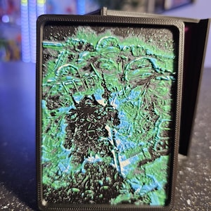 May include: A rectangular black case with a textured, colorful design. The design features a mix of green and blue hues, with a raised, three-dimensional appearance. The case is likely for a card or small item.