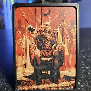 May include: A rectangular, black-framed pendant featuring a detailed illustration of a figure seated on a throne. The artwork is predominantly red and gold, depicting a regal scene with figures and spears.