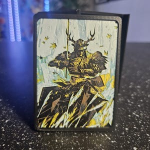 May include: A black card holder featuring a detailed illustration of a samurai warrior with antlers, holding a sword. The artwork uses a palette of black, gold, and white, set against a backdrop of stylized foliage.