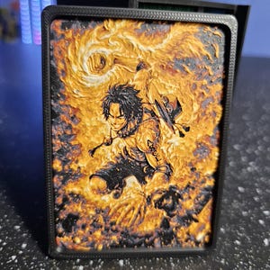 May include: A rectangular 3D printed plaque featuring a textured, multi-colored relief of a male anime character with spiky hair, a straw hat, and a tattoo on his arm, surrounded by swirling orange and yellow flames within a black frame.